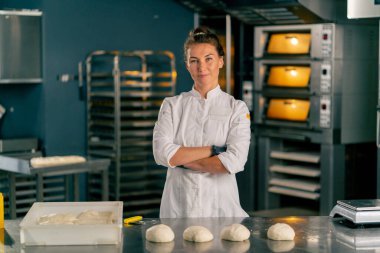 Kadın fırıncı, fırında pişirmek için hazırlanmış hamur ile profesyonel bir mutfak yüzeyinin arkasında duruyor ve kameraya bakıyor.