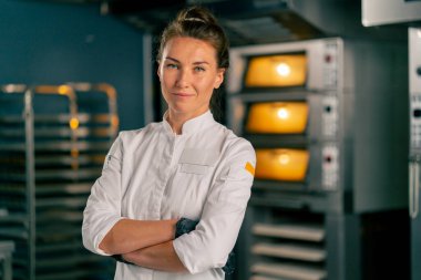 Kadın bir fırıncı, fırının arka planında profesyonel bir mutfak yüzeyinin arkasında duruyor ve kameraya bakıyor.