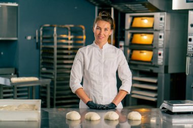 Kadın fırıncı, fırında pişirmek için hazırlanmış hamur ile profesyonel bir mutfak yüzeyinin arkasında duruyor ve kameraya bakıyor.