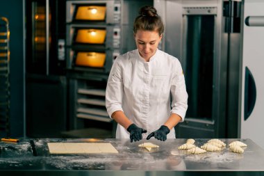 Ceketli ve eldivenli bir kız şef, bir fırında pişirmek için hamur hamurunun eşit parçalarından kruvasan yapar.