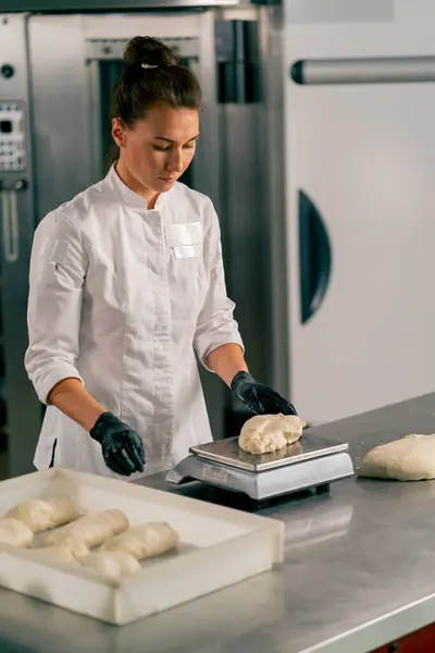 制服の女性パン屋は オーブンで焼く前にキッチン表面のクロワッサン形の生地を重量を量ります — ストック写真