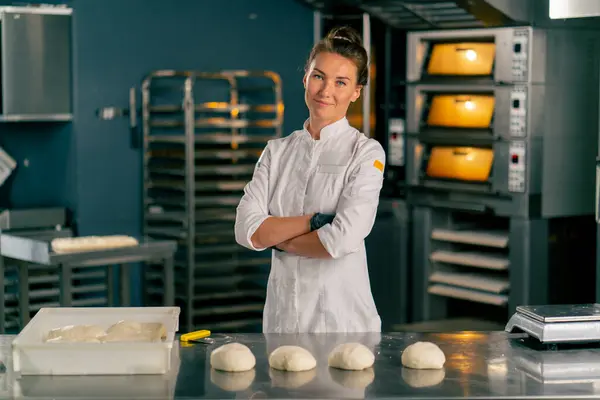 女面包师站在一个专业厨房表面的面包房后面 用准备好的面团烘焙 然后看着摄像机 — 图库照片