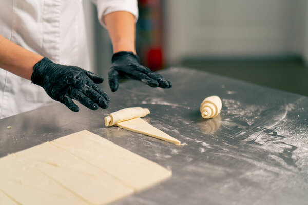 Close-up shot of a chef's hand in gloves rolling raw cut dough into croissant shapes for baking
