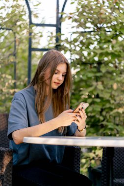 Üzgün genç bir kız bahçede bir masada oturuyor ve düşünceli bir şekilde elindeki telefona bakıyor.