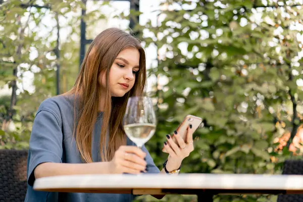 Sad young girl sitting at a table in the garden with a glass of wine and looking at the phone