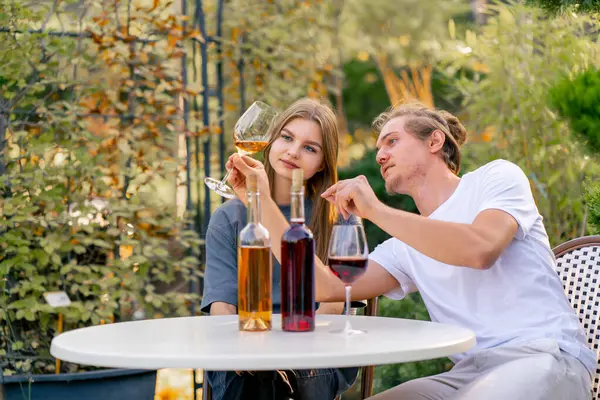 A couple of a guy and a girl are sitting together at a table in the garden of a winery and tasting different types of wine