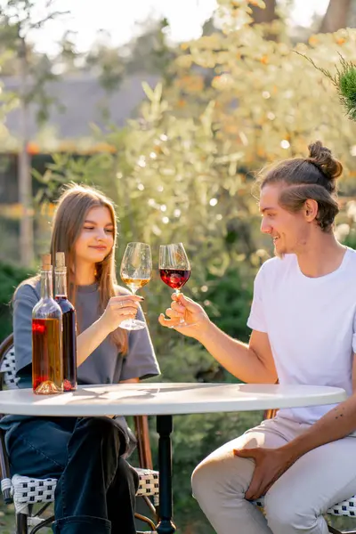 A couple of a guy and a girl are sitting together at a table in the garden of a winery clinking glasses of different types of wine