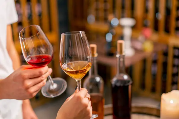 Close-up shot of a man and woman\'s hands holding glasses of different types of wine and clinking glasses at a tasting in the cellar