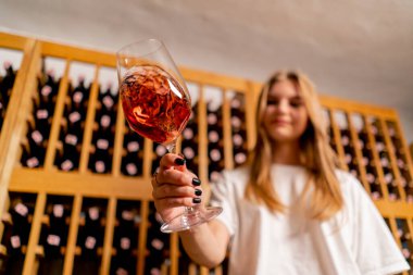 Bir kız garson kız şarap mahzeninde elinde bir bardak şarapla durup gülümseyerek kameraya uzatıyor.