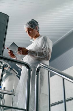 Beyaz koruyucu takım elbiseli ve başlıklı bir erkek teknoloji uzmanı bir bira fabrikasında içki üretim aşamasını kontrol ediyor.