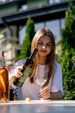 Genç ve güzel bir kız bir restoranın terasında otururken şişeden bardağa şarap dolduruyor.