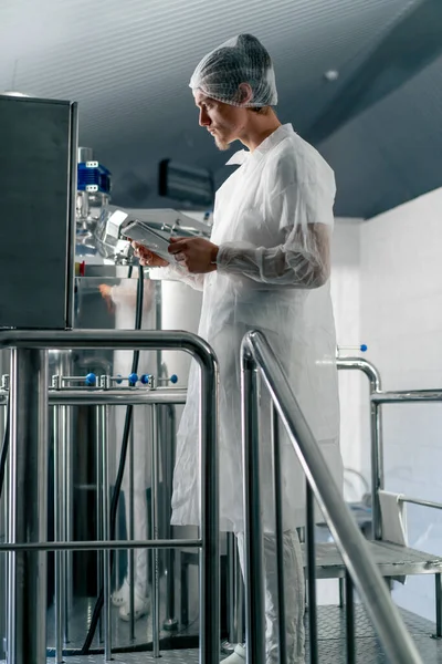 白い保護スーツとヘッジアの男性技術者は 醸造所で飲み物の生産段階をチェックする — ストック写真
