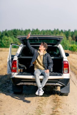 Araba orman bozkırında yolda duruyor. Genç bir adam açık bagajda telefonuyla oturuyor ve bağlantı kuruyor.
