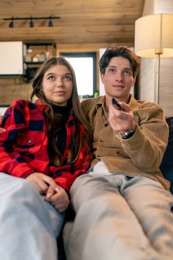 Birbirine aşık gülümseyen bir çift kanepede oturuyor ve rahat ahşap bir evde komedi filmi izliyorlar.
