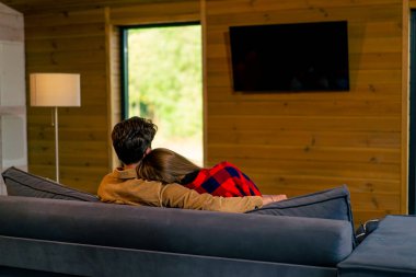 Genç bir çiftin evde oturup televizyon izlediği ve ormanın içinde güzel, ahşap bir evde sarılıp yattığı bir fotoğraf.