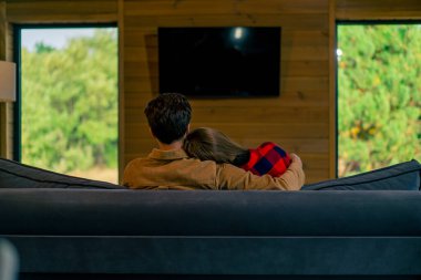 Genç bir çiftin evde oturup televizyon izlediği ve ormanın içinde güzel, ahşap bir evde sarılıp yattığı bir fotoğraf.