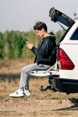 Genç bir adam elinde telefonla bir arabanın bagajında oturuyor ve çayırdaki bir durakta bölgeyi inceliyor.