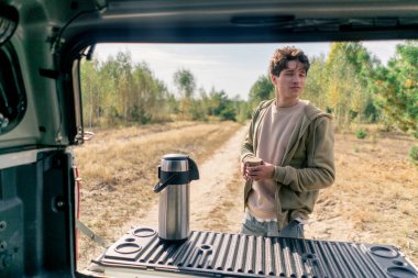 Adamın biri, büyük metal bir termostan sıcak çay içerken arabasının bagajını açıyor.