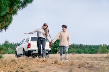 Genç bir çift, bir kız ve bir erkek el ele tutuşarak ve arabalarının arka planında konuşarak ormanda birlikte yürüyorlar.