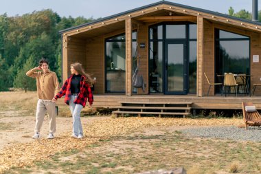 Bir adam ve bir kız, büyük panoramik pencereli ahşap evden el ele tutuşarak dışarı çıkarlar.