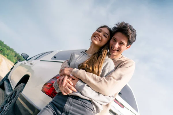 Portrait of a young woman and man laughing and hugging near their car while traveling on vacation