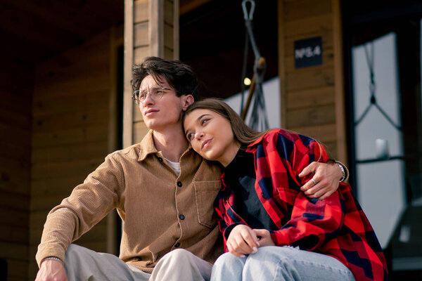 A young stylish couple in love is sitting together hugging on the steps of a large wooden cottage