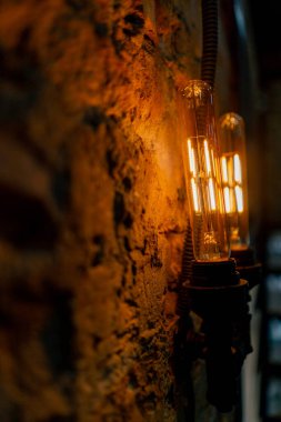 Alacakaranlıkta tavan arası tarzında tuğla bir duvarda parlayan kurşun ampullü duvar lambalarının yakın çekimi.