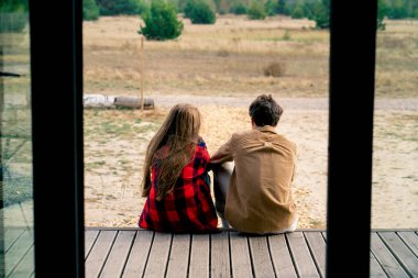 Bir adamla bir kızın, ahşap bir evin terasında oturup bozkırın ve ormanın doğasına hayran kaldıkları bir sahne.