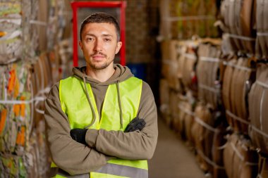 Koruyucu yelek giymiş ciddi bir adamın portresi kollarını önünde kavuşturmuş ve bir atık tesisine bakıyor.