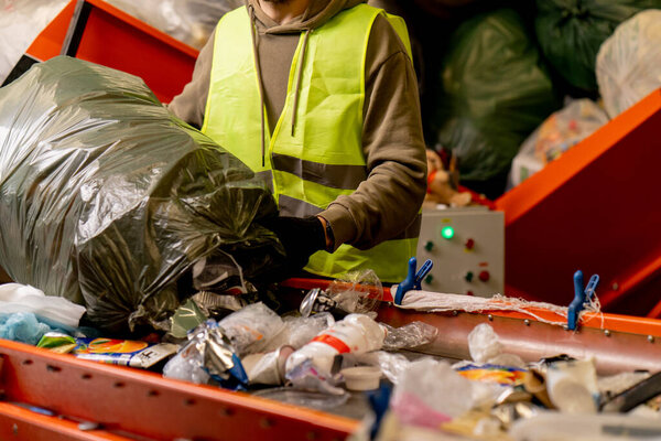 Focused male sorter with a protective vest and gloves unloads waste from a garbage bag onto a garbage sorting line