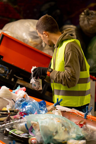 Side view of a sorter in gloves and a protective vest who sorts garbage on a special sorting line while working at a waste disposal station