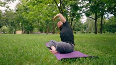 Yakışıklı sakallı adam parkta meditasyon yapıyor yoga yapıyor esneme hareketleri yapıyor ve farklı kas grupları çalıştırıyor.