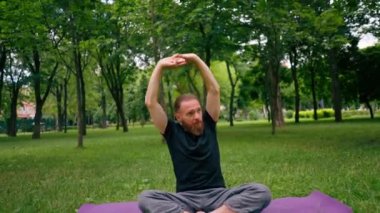Yakışıklı sakallı adam parkta meditasyon yapıyor yoga yapıyor esneme hareketleri yapıyor ve farklı kas grupları çalıştırıyor.