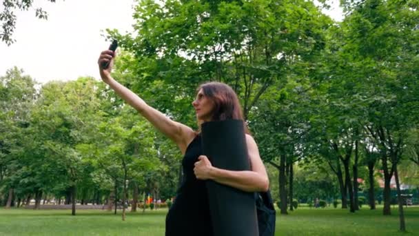 トレーニング前またはトレーニング後に公園で散歩中にアクティブな美しいヨギの女性は 彼女の手でフィットネスマットを保持する電話で写真を撮ります — ストック動画