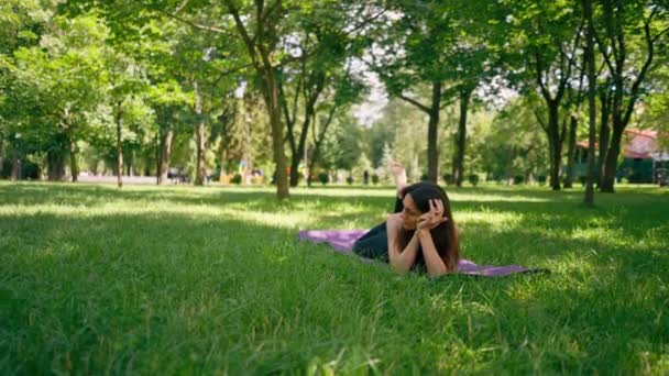 アクティブな美しいヨギの女性は 空を見ながらフィットネスマットに横たわった後 市内公園で休んでいます — ストック動画