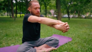Yakışıklı sakallı adam parkta meditasyon yapıyor yoga yapıyor esneme hareketleri yapıyor ve farklı kas grupları çalıştırıyor.