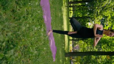 Deneyimli bir kadın eğitmen parkta meditasyon yaparken dikey videoda yoga yapıyor ve farklı kas grupları uyguluyor.