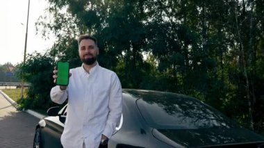 Siyah lüks bir arabanın yanında duran gülümseyen sakallı adam yeşil ekranlı akıllı bir telefon ve kiralık bir araba gösteriyor.