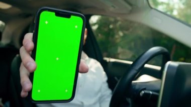 Yakın plan gülümseyen sakallı iş adamı. Lüks araba kullanırken yeşil ekranlı telefonu gösteriyor.