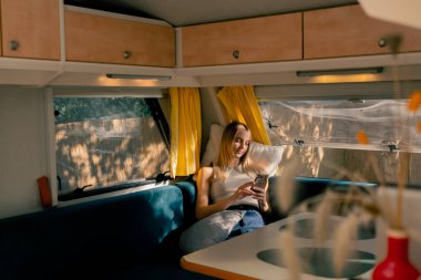 Yorgun bir kız pencerenin yanındaki karavanda kanepede oturur ve seyahat ederken dikkatlice telefonuna bakar.