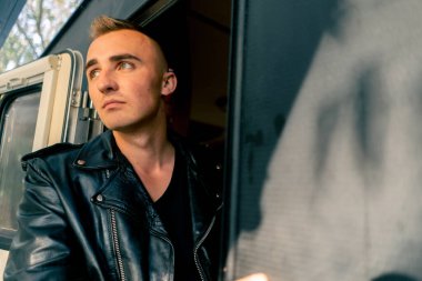 Deri ceketli şık bir adam seyyar bir evden terasa çıkar ve çevreye bakar.