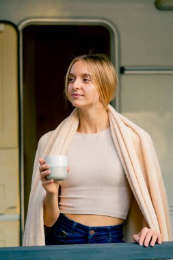 Omuzlarında battaniyesi olan genç bir kız terasta sabah kahvesini içiyor.