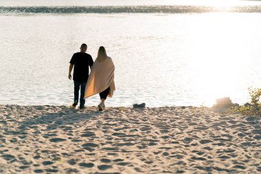 Birbirine aşık çiftler battaniyeyle sarılıp gün batımında kumlu sahil boyunca nehre doğru yürürler.