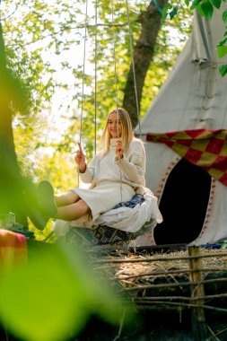 Güzel elbiseli genç bir kız ağaçların arkasında salıncakta sallanıyor ve gülümsüyor.