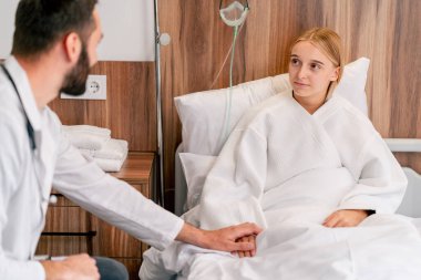 Doktor hasta kıza bir hastane odasında destek oluyor ve iyileşmesi için elini tutuyor.