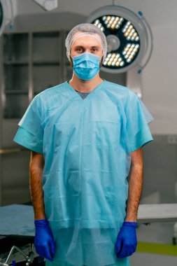 Üniformalı ve koruyucu şapkalı ciddi bir erkek cerrah ellerini önünde kavuşturuyor ve ameliyathanede kameraya bakıyor.