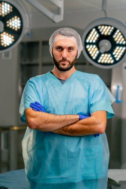 Üniformalı ve koruyucu şapkalı ciddi bir erkek cerrah ellerini önünde kavuşturuyor ve ameliyathanede kameraya bakıyor.