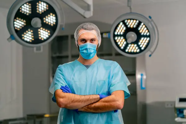Serious male surgeon in uniform and protective cap folds his hands in a front of him and looks at the camera in operating room