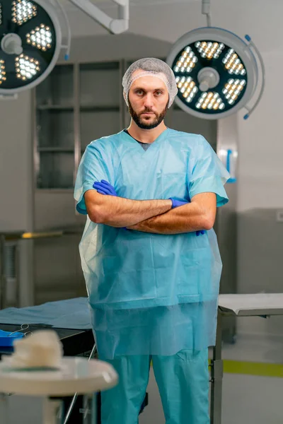 Üniformalı ve koruyucu şapkalı ciddi bir erkek cerrah ellerini önünde kavuşturuyor ve ameliyathanede kameraya bakıyor.