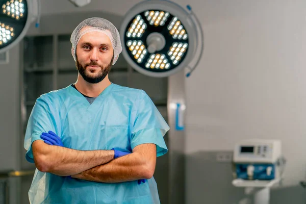 Serious male surgeon in uniform and protective cap folds his hands in a front of him and looks at the camera in operating room